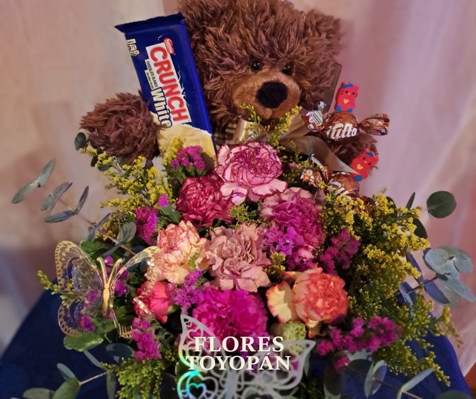 Arreglo floral que contiene un peluche de oso y chocolate